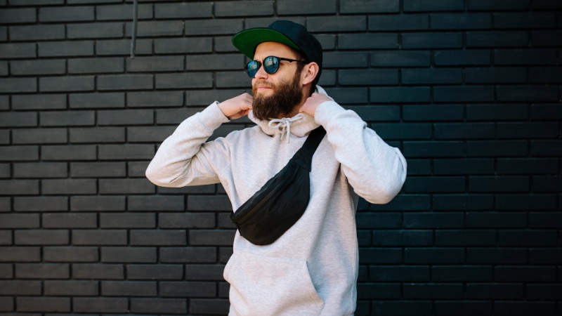Mann in minimalistisch/funktionalem Style, vor einer Wand mit Bauchtasche, Hoodie, Cap und Sonnenbrille - sieht aus wie Mark Forster