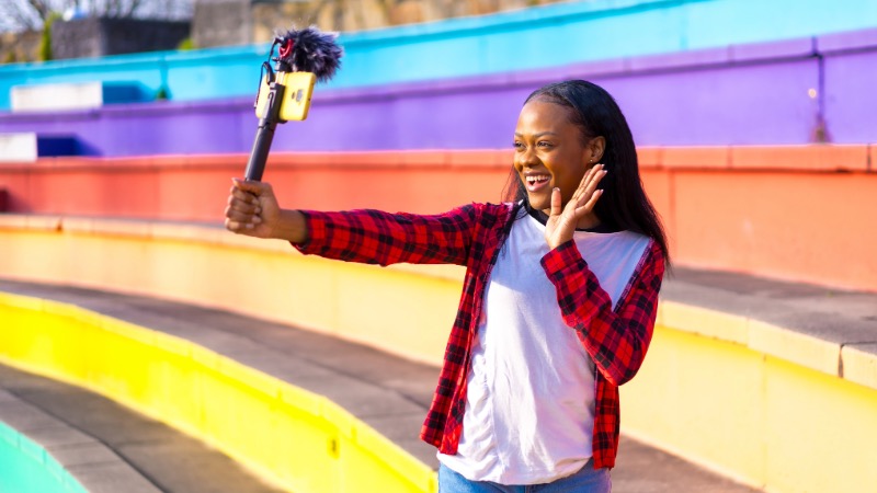 Frau vor farbenfrohem Hintergrund, redet mit ihrem mobilen Endgerät, dieses ist verbunden mit einem Mikrofon sowie einem Stick für professionellere Influencer-Aufnahmen